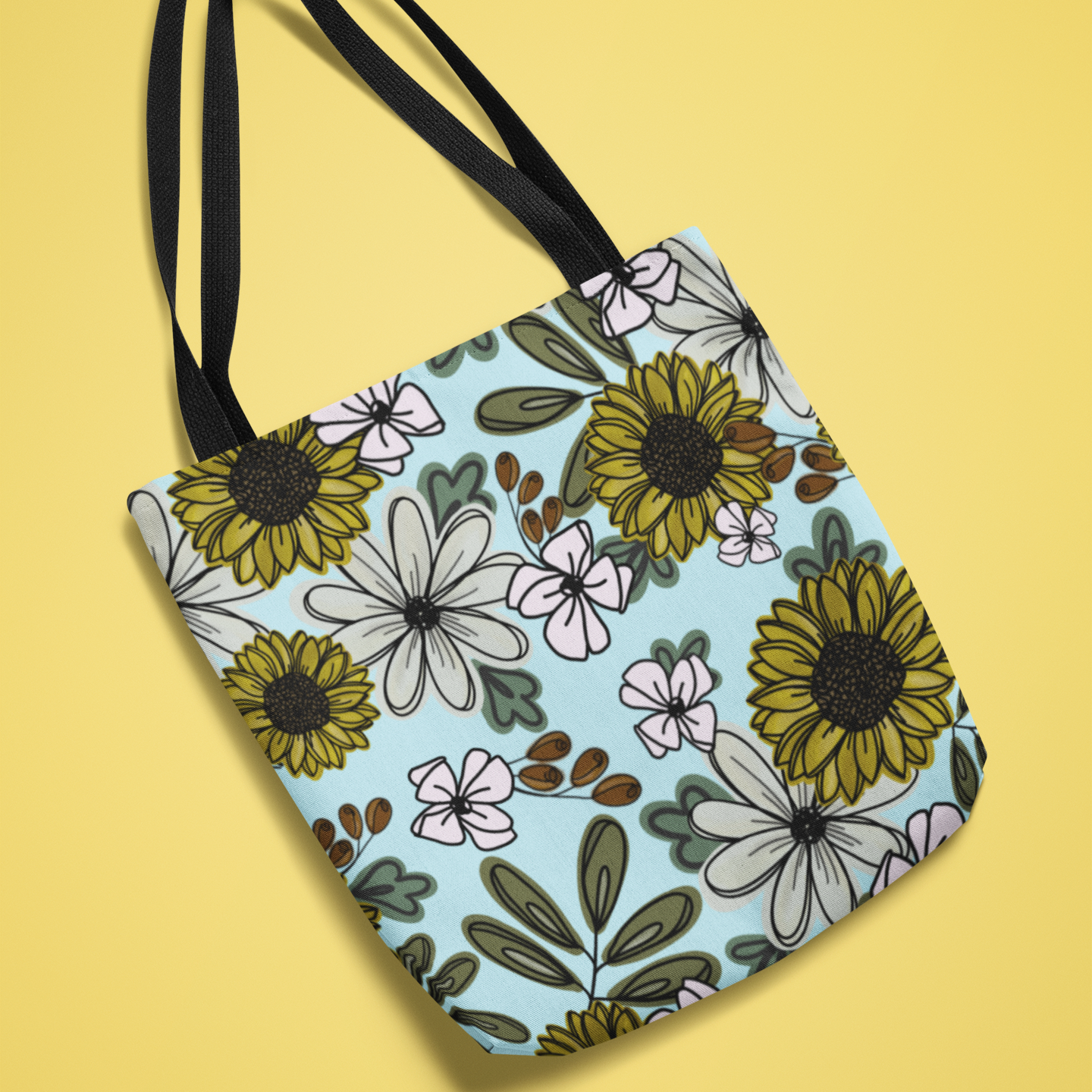 Canvas tote bag with a vibrant sunflower and wildflower pattern on a light blue background, displayed against a yellow backdrop. Features black handles and a spacious design, perfect for shopping or everyday use