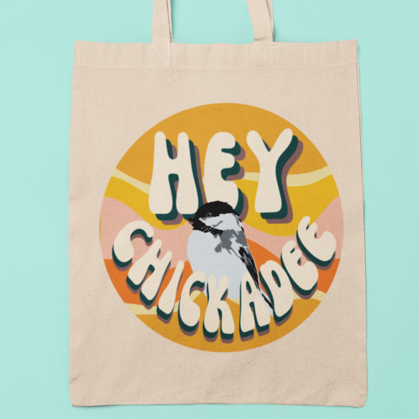 Natural canvas tote bag featuring the retro 'Hey Chickadee' design with vibrant typography and a chickadee illustration, displayed against a bright blue background.