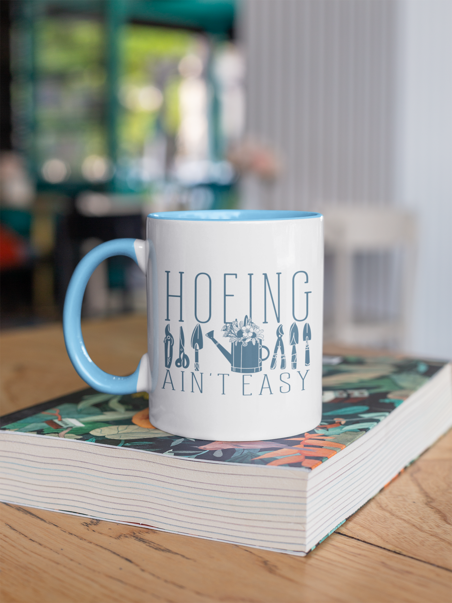 White ceramic mug with 'Hoeing Ain't Easy' text and garden tool illustrations, styled on a desk with books and plants for a cozy and creative setup.