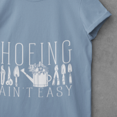 Close-up of the 'Hoeing Ain't Easy' design on a slate blue t-shirt, highlighting the bold typography and detailed gardening tool illustrations. Perfect for adding humor to a gardener's wardrobe.