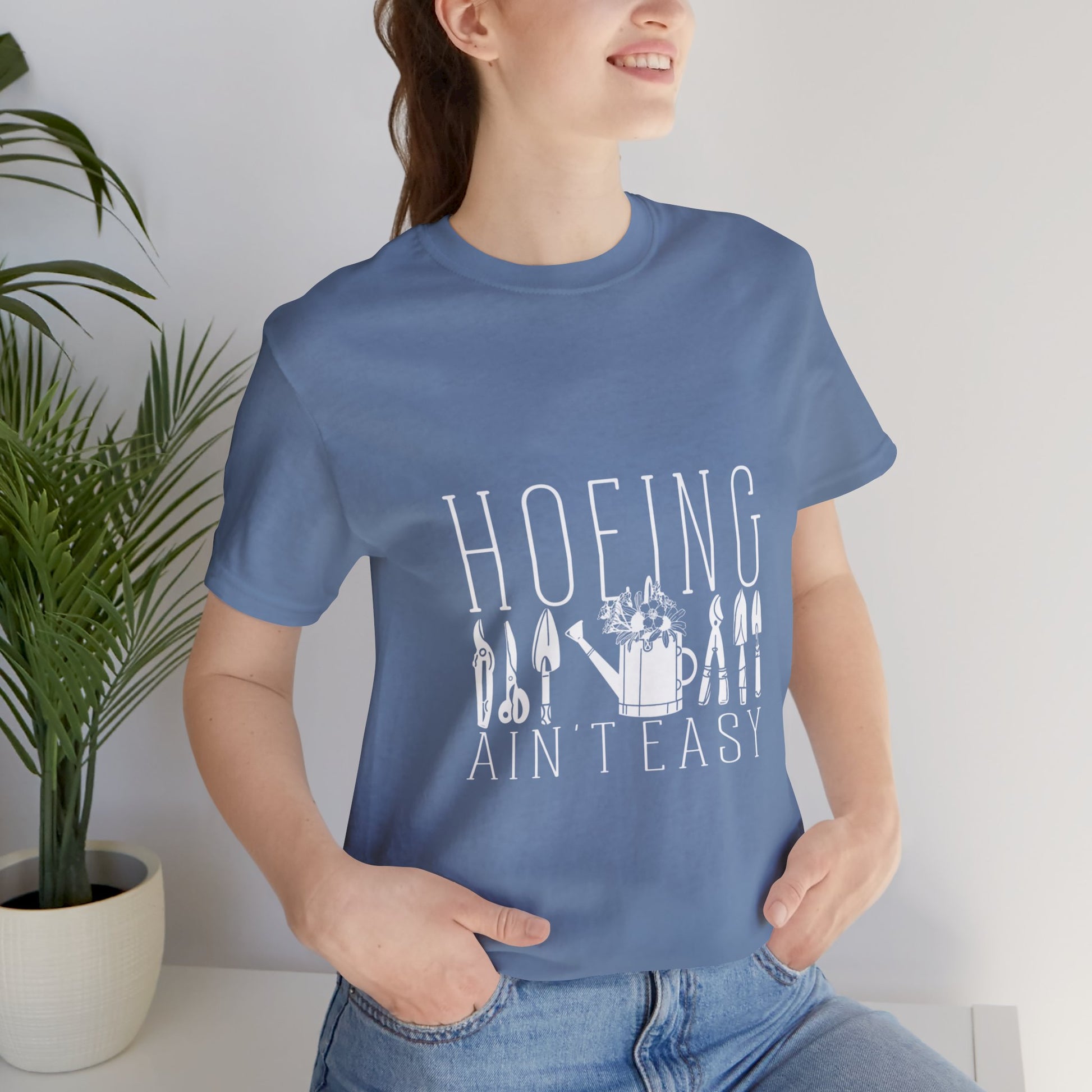 Funny gardening t-shirt with 'Hoeing Ain't Easy' design in slate blue, worn by a person outdoors in a garden setting. Perfect for nature lovers and gardeners