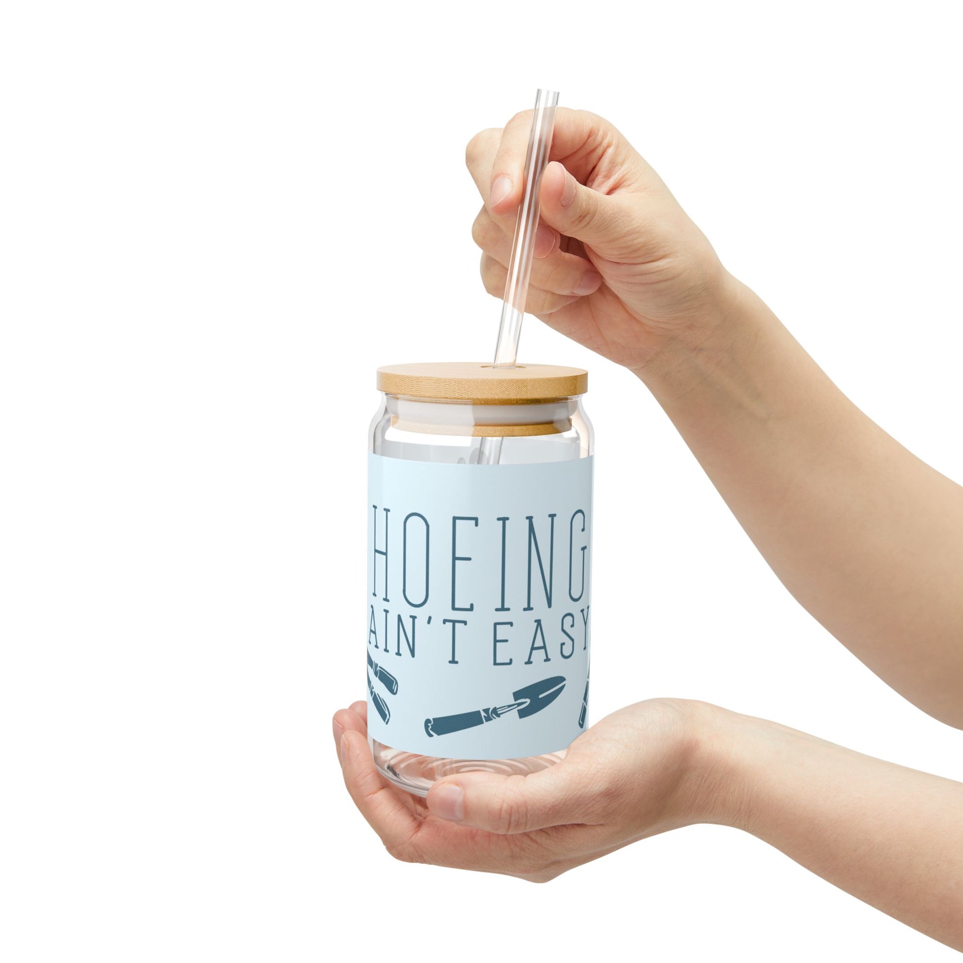 Reusable glass tumbler with 'Hoeing Ain't Easy' gardening design, held in hand to showcase its size and ergonomic fit. Features a bamboo lid and straw.