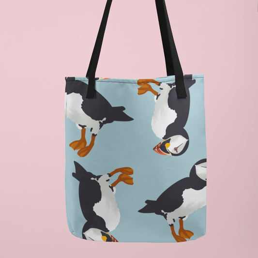 Canvas tote bag featuring a whimsical puffin bird pattern on a light blue background with black handles, displayed against a pastel pink backdrop. Perfect for bird lovers and nature enthusiasts.