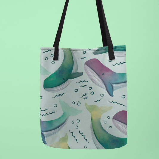 Canvas tote bag featuring a whimsical watercolor whale pattern with splashes of blue and purple hues, displayed against a soft green background. Includes durable black handles.